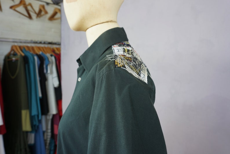 May include: A dark green long-sleeved shirt with a decorative patch on the shoulder. The patch is made of various fabrics and includes a small label with the text "31".