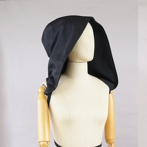 Può includere: Un cappuccio nero in tessuto, esposto su un manichino. Il cappuccio è drappeggiato sulla testa e sulle spalle del manichino, con il tessuto che cade in morbide pieghe. Il design del cappuccio è semplice ed elegante, adatto a vari usi di costume o moda.