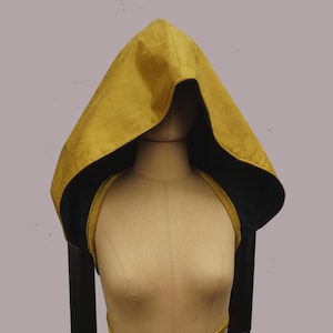 May include: A mustard yellow hooded cloak with a black lining, shown on a mannequin. The cloak features a pointed hood and is draped over the shoulders. The garment is made of a soft, textured fabric.
