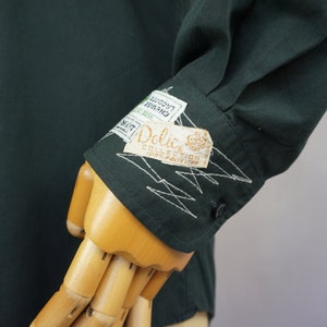 May include: A dark green long-sleeved shirt with a white embroidered design on the cuff. The shirt has a label that reads "Delic. Collection 100% Pavlova 2"