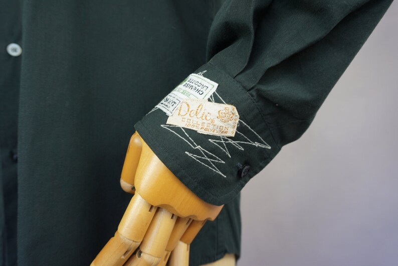 May include: A dark green long-sleeved shirt with a white embroidered design on the cuff. The shirt has a label that reads "Delic. Collection 100% Pavlova 2"