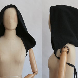 May include: Black hooded cowl, shown on a mannequin. The cowl is made of a textured fabric with a wide hood and shoulder coverage. It has ties for an adjustable fit. The background is plain white.