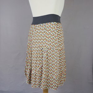 Może przedstawiać: Spódnica w szaro-biało-pomarańczowe wzory z czarnym pasem. Spódnica wykonana jest z miękkiej, przewiewnej tkaniny i ma warstwowy krój.