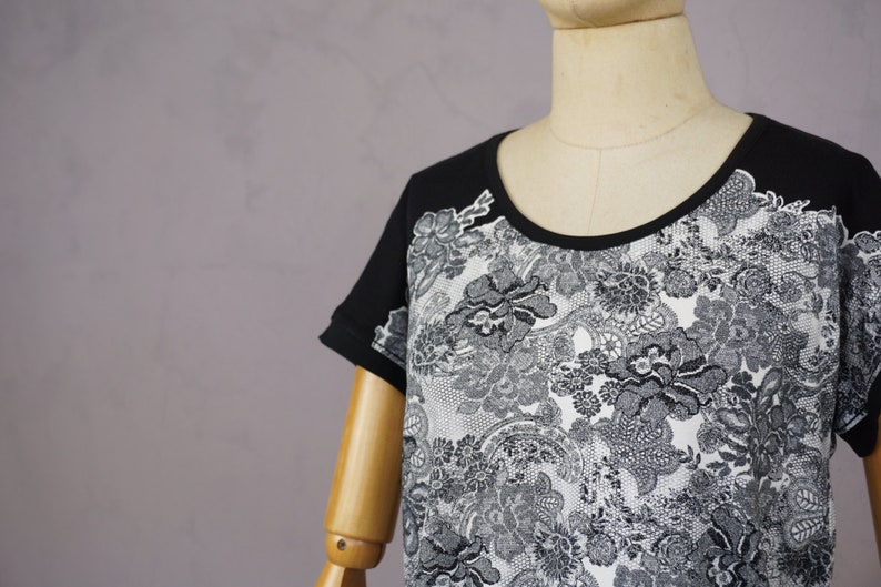 May include: Black and white short-sleeved shirt with a lace pattern. The shirt has a round neckline and is made of a soft, comfortable fabric.