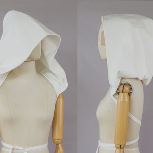May include: A white hooded garment, possibly a cloak or cowl, displayed on a mannequin. The hood is large and draped, with a structured shape. The garment has a shoulder covering and ties at the waist.