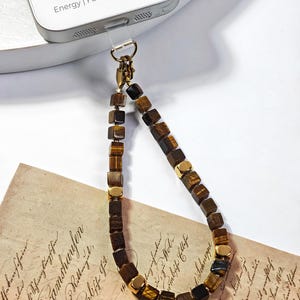 May include: A phone charm strap made of cube-shaped brown tiger's eye beads and small gold-toned metal spacers. The strap is attached to a silver-colored smartphone with the text Energy | Focus | Confidence printed on the back.