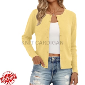 May include: A light yellow knit cardigan featuring a round neckline, long sleeves, and a front button closure. The garment is worn over a white top and blue denim shorts. The text KNIT CARDIGAN is visible in the center of the image.