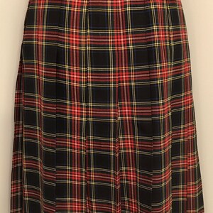 May include: A black, red, yellow, and white plaid pleated skirt. The skirt has a waistband with a button closure and is displayed on a mannequin. The fabric appears to be a medium-weight material. The skirt is a classic design.