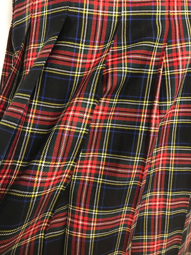 May include: A close-up of a pleated tartan fabric. The pattern is a classic plaid design, with a black background and intersecting lines in red, yellow, white, and blue. The fabric is folded, showing the texture and drape of the material.