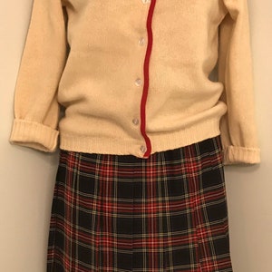 May include: A cream-coloured cardigan with red trim and embroidered red and blue cross-stitch details. It is paired with a black, red, and white plaid pleated skirt. A vintage outfit.
