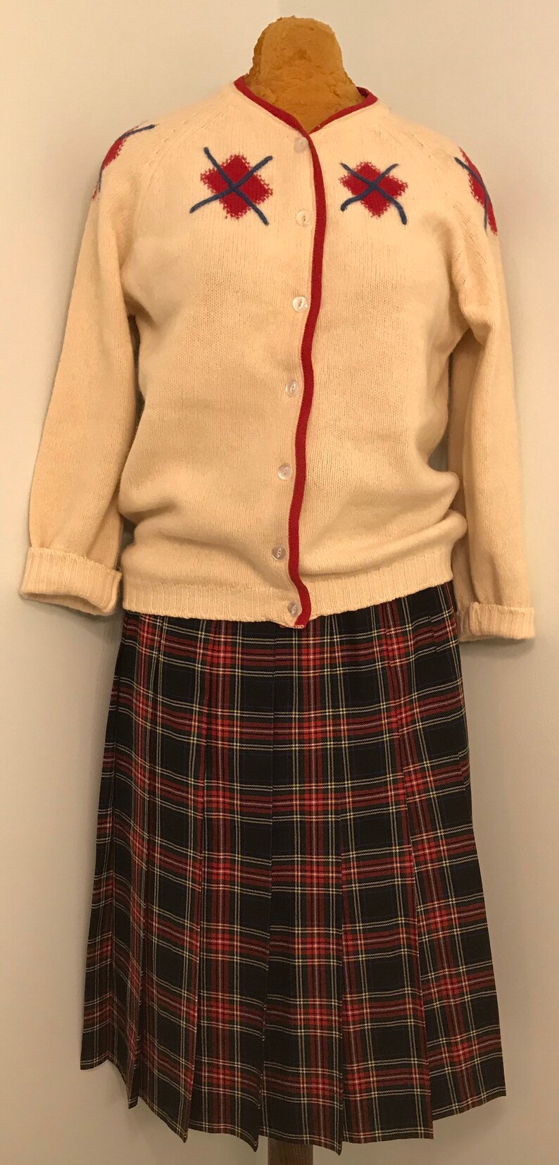 May include: A cream-coloured cardigan with red trim and embroidered red and blue cross-stitch details. It is paired with a black, red, and white plaid pleated skirt. A vintage outfit.