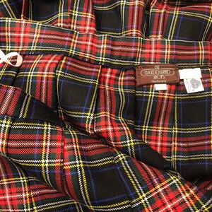 May include: Close-up of a pleated plaid skirt in red, black, green, yellow, and blue. The skirt has a white ribbon detail and a brown tag that reads "The Woodward Shops". The fabric is folded and draped.
