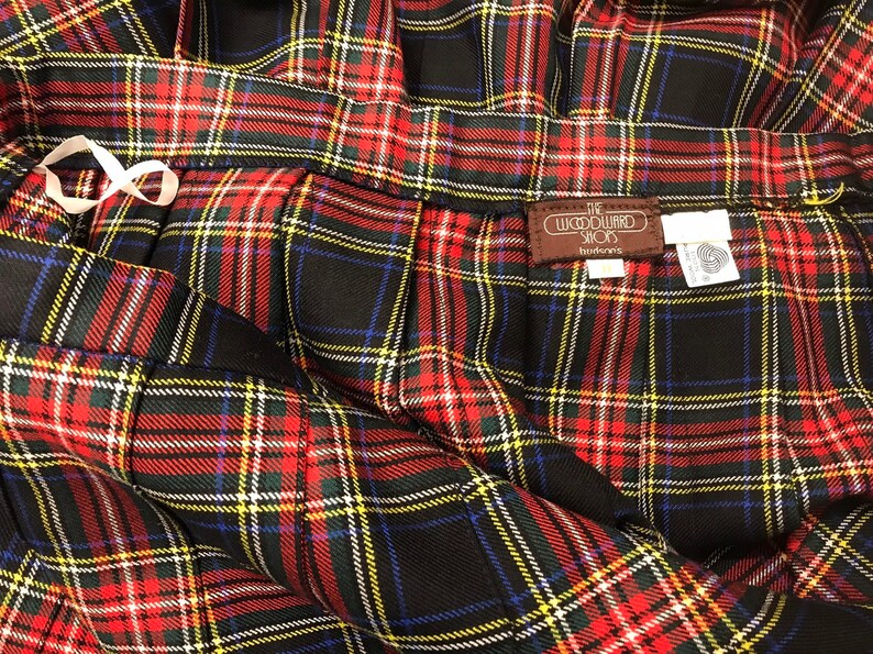 May include: Close-up of a pleated plaid skirt in red, black, green, yellow, and blue. The skirt has a white ribbon detail and a brown tag that reads "The Woodward Shops". The fabric is folded and draped.