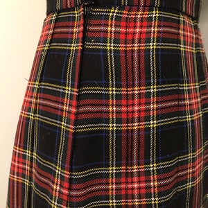 May include: Close-up of a black tartan skirt featuring red, yellow, blue, and white lines. The skirt has a zip closure and a waistband. The fabric is woven, and the skirt appears to be pleated.