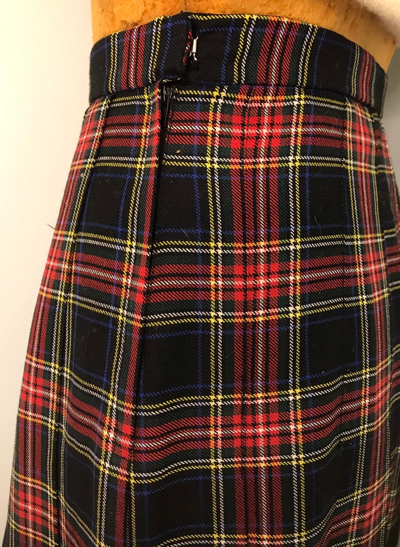 May include: Close-up of a black tartan skirt featuring red, yellow, blue, and white lines. The skirt has a zip closure and a waistband. The fabric is woven, and the skirt appears to be pleated.