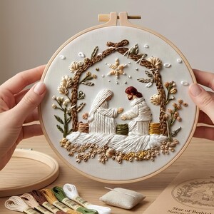 May include: A circular wooden embroidery hoop containing a white fabric panel with a hand-stitched nativity scene. The design features two figures in white robes under a brown arched structure, a star above, and floral accents. Embroidery floss and a needle cushion are nearby.