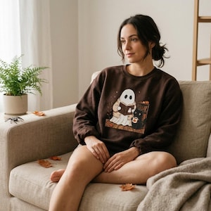 Può includere: Una felpa girocollo marrone scuro con un'illustrazione grafica di un fantasma bianco che indossa un cardigan beige e legge un libro, accompagnato da un gatto nero che tiene una tazza, su uno sfondo di foglie autunnali e una libreria.