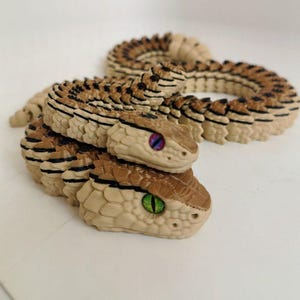 Könnte beinhalten: Ein zweiköpfiges, bewegliches Schlangenspielzeug mit einem beigefarbenen und braunen Schuppenmuster sowie schwarzen Akzenten. Der obere Kopf hat violette Augen, der untere Kopf hat grüne Augen. Der Körper ist segmentiert und flexibel beweglich.