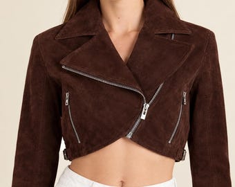 May include: A cropped dark brown suede jacket featuring a wide notched collar, an asymmetrical silver-tone metal zipper closure, and two diagonal zippered pockets on the front. The jacket includes small side buckles at the hemline.