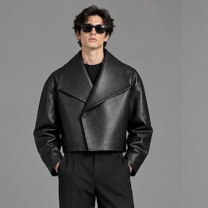 May include: A black cropped leather jacket with wide lapels and long sleeves, worn over a black shirt and black trousers. The jacket features a smooth texture and a structured, boxy fit. Dark sunglasses are worn on the face.