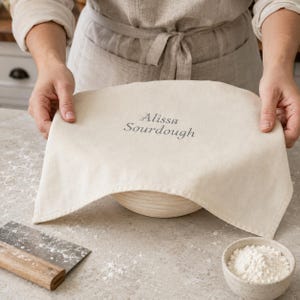 May include: A cream-coloured fabric proofing cloth covering a round sourdough banneton bowl. The cloth features the embroidered text Alissa Sourdough in a dark grey thread. The fabric is draped over the bowl, showing a simple hemmed edge.