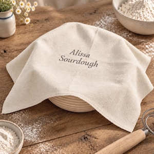 May include: A square, off-white cotton proofing cloth draped over a round, ridged rattan banneton basket. The fabric features the embroidered text Alissa Sourdough in a dark gray thread centered on the top surface of the cloth.