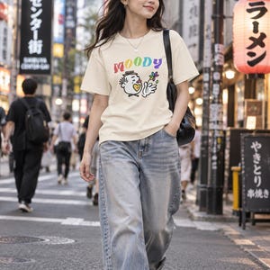 May include: A cream-colored short-sleeve t-shirt featuring the word MOODY in rainbow-colored letters above a cartoon character graphic. The character has a yellow heart on its chest, a small hat, and is holding a colorful flower. The shirt is paired with blue denim jeans and a black shoulder bag.