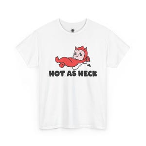 May include: A white short-sleeve t-shirt featuring a graphic of a red cartoon devil character lying down. Below the character, the text "HOT AS HECK" is printed in bold black letters. The shirt has a round neckline and a small circular logo on the inner neck.