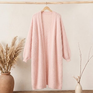 May include: A long-sleeved, open-front cardigan in a soft pastel pink color. The garment features a fuzzy, textured knit fabric, a ribbed collar, and ribbed cuffs at the end of the sleeves. It hangs straight down in a relaxed, knee-length silhouette.