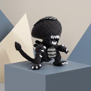 May include: A black and grey crocheted creature doll featuring a large elongated head, a wide open mouth with white teeth, sharp claws on its hands and feet, and a long pointed tail. The yarn work displays a textured pattern throughout the body.