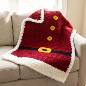 May include: A red crochet blanket featuring a white fuzzy border, a horizontal black stripe across the middle, a yellow rectangular belt buckle, and three circular yellow buttons arranged vertically along the upper portion of the blanket.