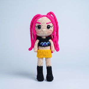 May include: A handmade crochet doll featuring bright pink yarn hair, a black crop top with a blue and purple mountain graphic, a yellow skirt, and black boots. The doll has a beige complexion, embroidered facial features, and pink blush cheeks.
