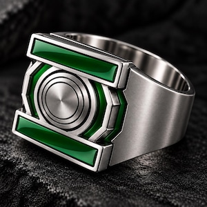 Peut inclure: Une bague en métal argenté présentant une face rectangulaire proéminente avec un emblème circulaire central. Le design comprend deux barres horizontales en émail vert entourant le centre, avec une finition métallique brossée sur l'anneau.