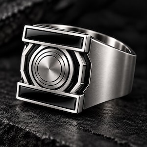 Könnte beinhalten: Ein Ring aus silberfarbenem Metall mit einem zentralen kreisförmigen Emblem mit konzentrischen Ringen. Das Design umfasst zwei horizontale schwarze rechteckige Einlagen über und unter der Mitte, eingefasst in einen geometrischen Rahmen.