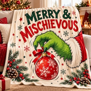 May include: A cream-colored throw blanket featuring the text MERRY & MISCHIEVOUS in green and red. A green furry hand with a red and white cuff holds a red Christmas ornament with white snowflakes. Pine branches and pinecones decorate the corners.