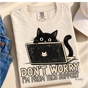 May include: An ivory t-shirt featuring a graphic of a black cat sitting behind a laptop. The laptop has a small white cat icon on its lid. Below the graphic, the text reads: DON'T WORRY I'M FROM TECH SUPPORT. The shirt has a Comfort Colors label.