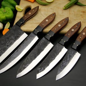 Pode incluir: Um conjunto de cinco facas de cozinha com lâminas de metal forjado de textura escura e cabos de madeira polida. Cada cabo apresenta uma incrustação circular distinta e rebites de metal. As facas estão dispostas em uma fileira.