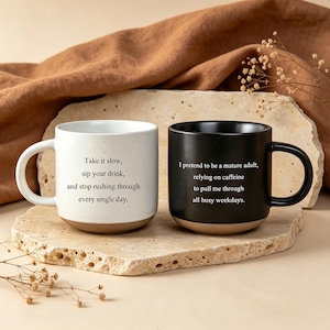 Puede incluir: Una taza de cerámica blanca con textura moteada y texto negro Take it slow, sip your drink, and stop rushing through every single day, junto a una taza negra mate con texto blanco I pretend to be a mature adult, relying on caffeine to pull me through all busy weekdays.