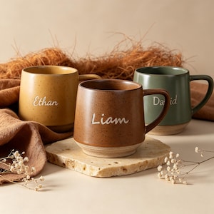 May include: Three ceramic mugs with speckled finishes in mustard yellow, terracotta brown, and sage green. Each mug features a handle and a personalized name engraved in white script: Ethan on the yellow mug, Liam on the brown, and David on the green.