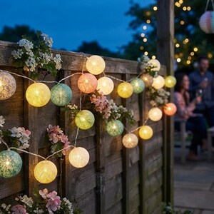 Puede incluir: Una guirnalda de luces LED esféricas con pantallas de tela texturizada en tonos amarillos, verdes, rosas y blancos. Los globos iluminados están dispuestos a lo largo de un cable fino, intercalados con flores artificiales de color rosa y blanco.