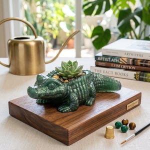 May include: A dark green, textured alligator-shaped planter with a succulent plant nestled in its back, resting on a rectangular wooden base. The base features a small gold-coloured plaque, accompanied by a gold thimble, four small beads, and a paintbrush.