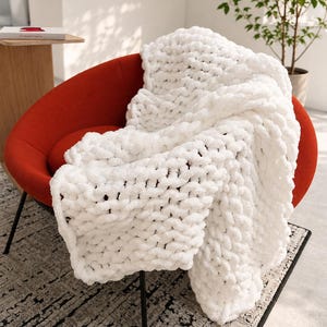 May include: A chunky knit throw blanket in a bright white color with a thick, looped texture. The blanket is draped over a curved, vibrant orange chair with thin black metal legs, showcasing the soft, voluminous yarn construction.