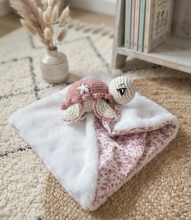 Peut inclure: Une couverture de sécurité en crochet en forme de tortue avec une tête crème aux yeux noirs brodés, une carapace rose poudré ornée d'une fleur blanche, et un dessous en peluche blanche douce associé à un tissu à motif floral rose et blanc.
