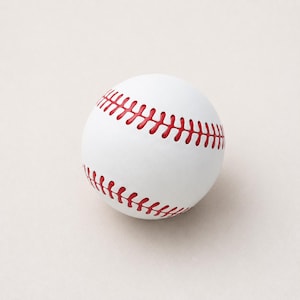 Könnte beinhalten: Ein weißer Baseball mit markanten roten Nähten, die sich über die Oberfläche der Kugel ziehen. Der Ball hat eine glatte, matte Textur und eine klassische runde Form, die das ausgeprägte Muster der roten Fadennaht deutlich hervorhebt.