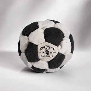 May include: A plush ball constructed from alternating black and off-white fabric panels. One off-white panel features black text reading SOUTHERN UTAH BASEBALL, EST. 2019, and a central SD logo flanked by two small stars.