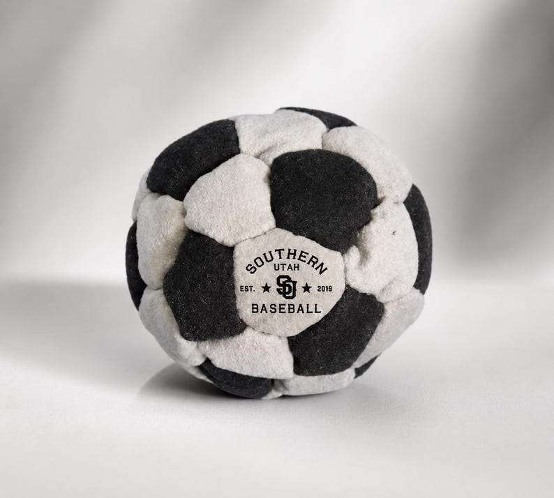 K&ouml;nnte beinhalten: Ein Pl&uuml;schball aus abwechselnden schwarzen und cremewei&szlig;en Stoffbahnen. Auf einer cremewei&szlig;en Bahn befindet sich der schwarze Schriftzug SOUTHERN UTAH BASEBALL, EST. 2019 sowie ein zentrales SD-Logo zwischen zwei kleinen Sternen.