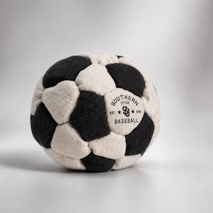 K&ouml;nnte beinhalten: Ein kugelf&ouml;rmiger Ball aus abwechselnden schwarzen und cremefarbenen Filzstoffbahnen. Ein cremefarbenes Feld zeigt ein schwarzes aufgedrucktes Logo mit der Aufschrift SOUTHERN UTAH BASEBALL, den Buchstaben SU und dem Text EST. 2016.