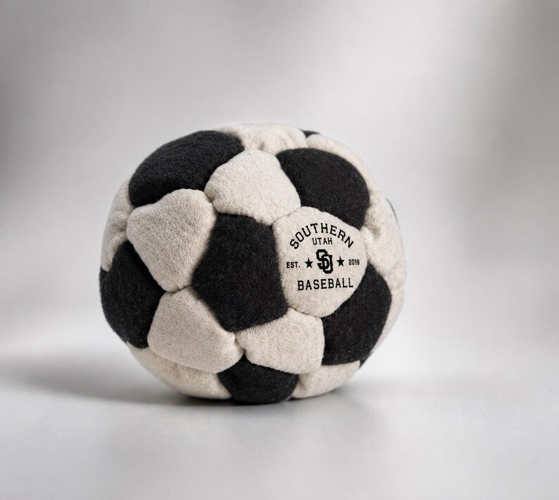 K&ouml;nnte beinhalten: Ein kugelf&ouml;rmiger Ball aus abwechselnden schwarzen und cremefarbenen Filzstoffbahnen. Ein cremefarbenes Feld zeigt ein schwarzes aufgedrucktes Logo mit der Aufschrift SOUTHERN UTAH BASEBALL, den Buchstaben SU und dem Text EST. 2016.