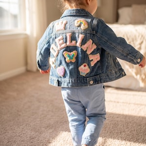 Peut inclure: Une veste en jean bleu avec des écussons brodés au dos. Les motifs incluent le nom ADELINA en lettres roses, une couronne, un arc-en-ciel, un papillon, un cœur et un cupcake. La veste possède un col, des manches longues et des boutons en métal.
