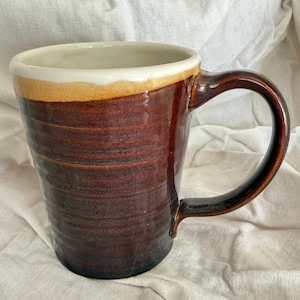 May include: A ceramic mug with a dark brown, ribbed exterior and a smooth, cream-colored rim. The mug features a glossy finish, a wide opening, and a curved handle on the right side. The body has a cylindrical shape with subtle horizontal ridges.